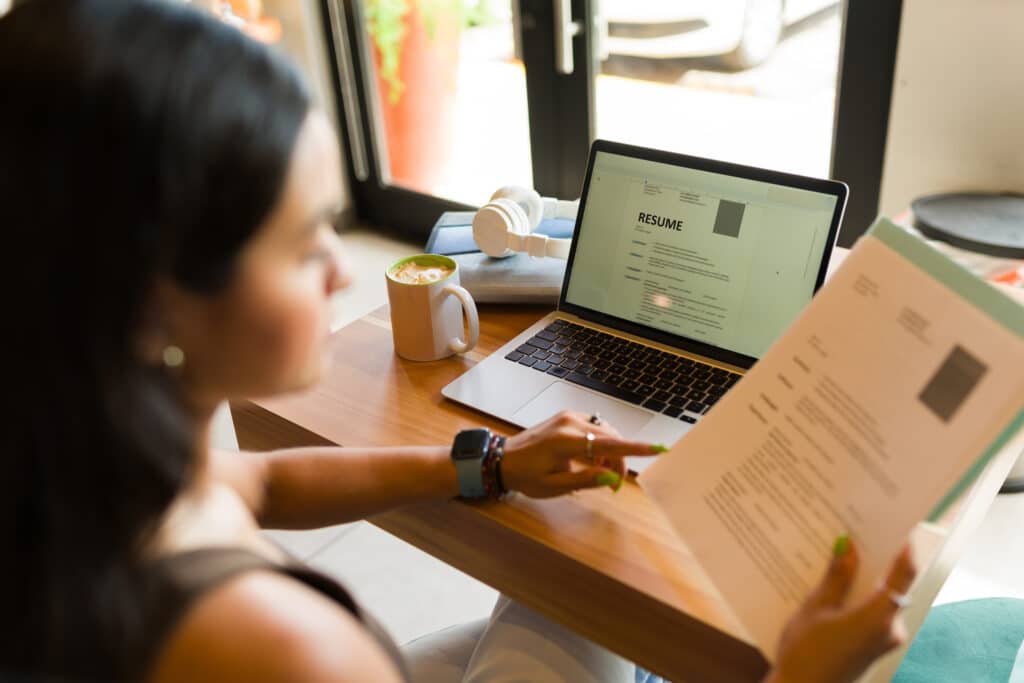Person sits at a desk reviewing a resume on a laptop, with a mug of coffee and headphones nearby.