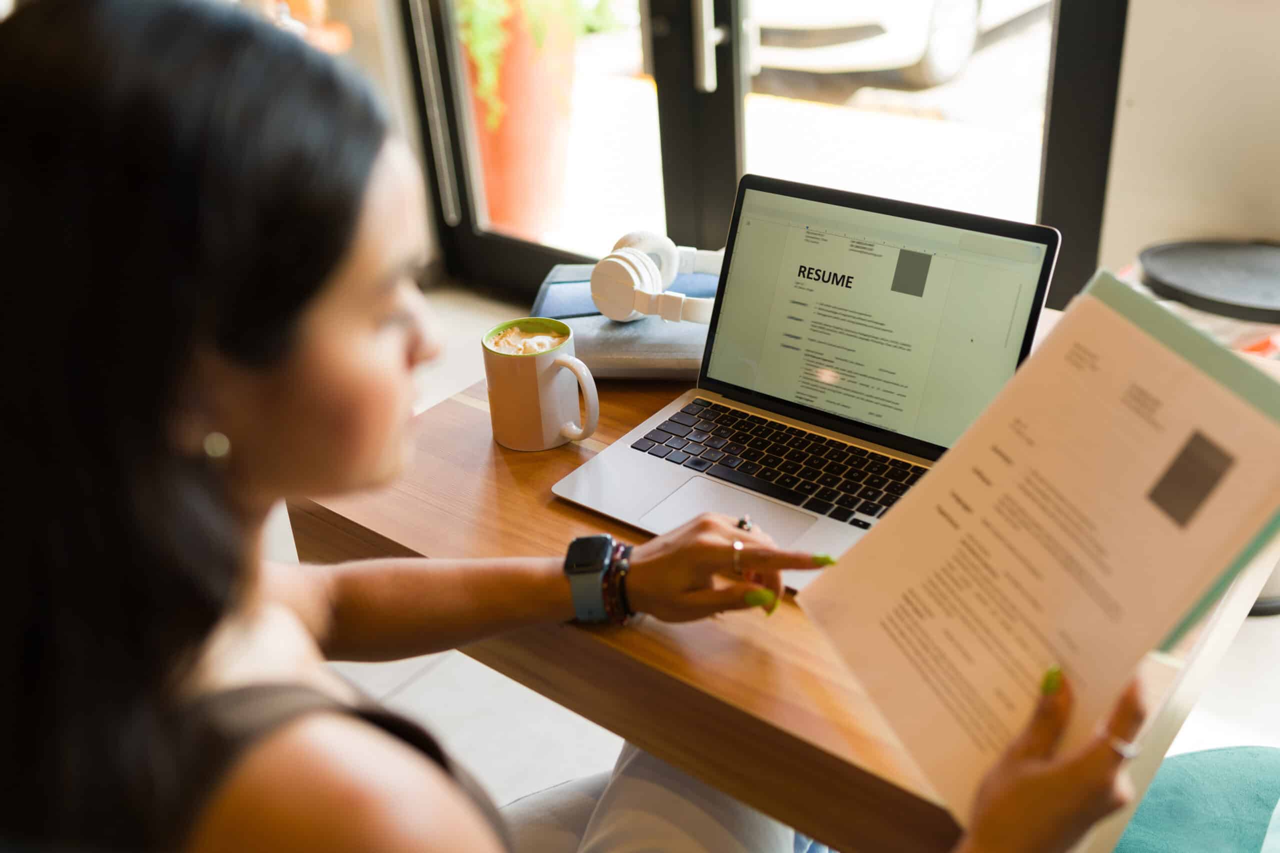 Person sits at a desk reviewing a resume on a laptop, with a mug of coffee and headphones nearby.