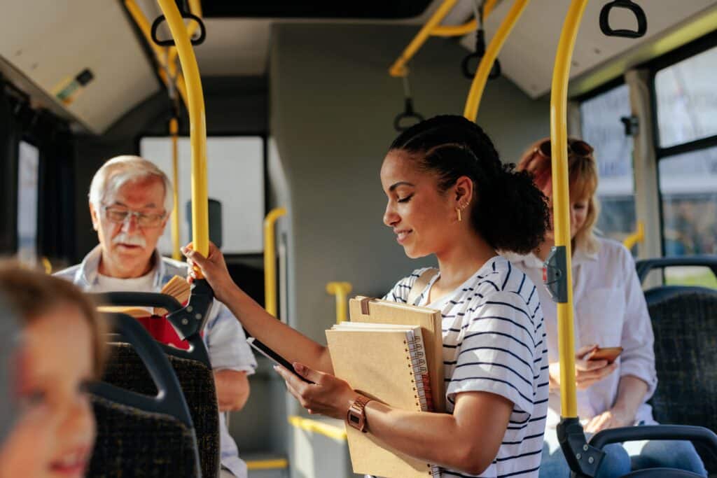 Young woman standing on a bus, holding a clipboard and notebook, looking at her phone while others sit nearby.