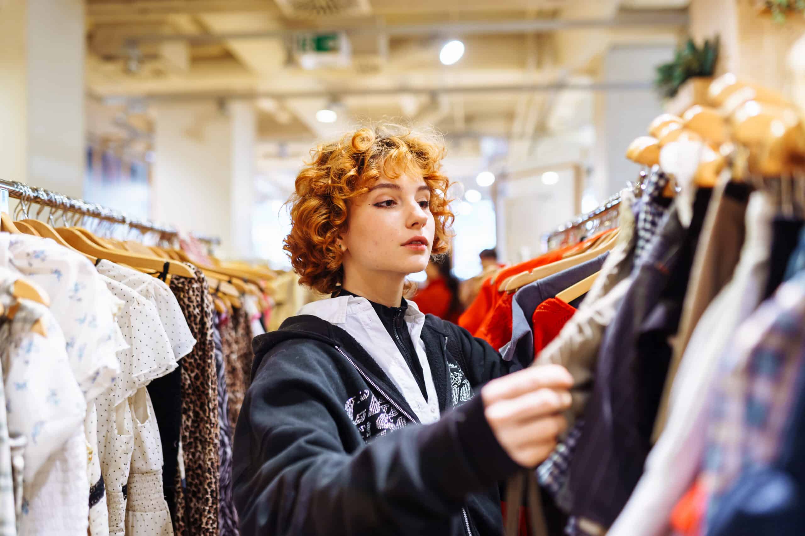 Woman with curly red hair browsing clothes on a rack in a store in a bright boutique setting.